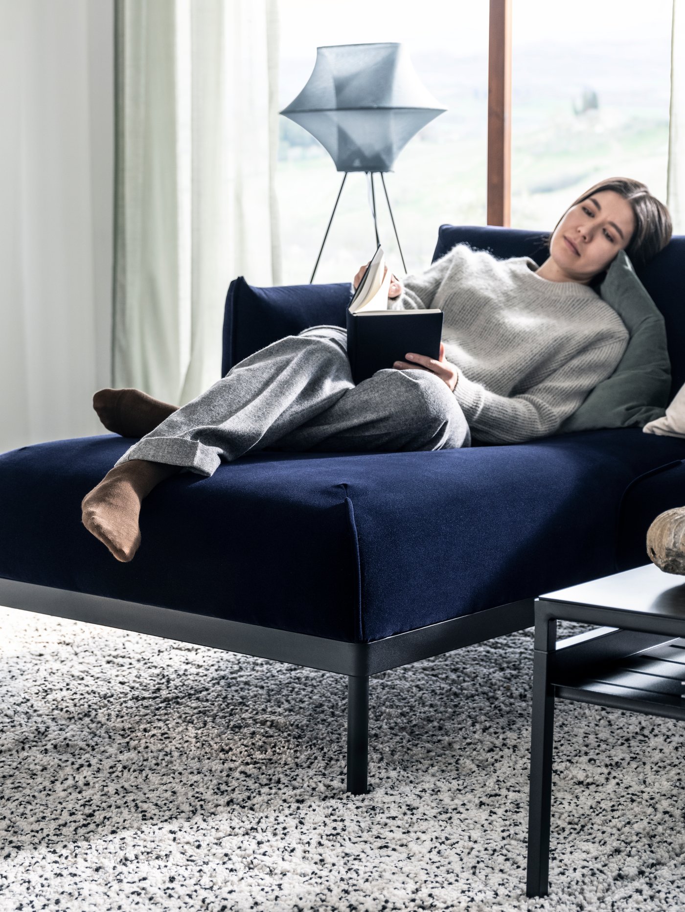 A woman in grey loungewear reading a book and reclining on a dark blue ÄPPLARYD sofa with chaise longue.