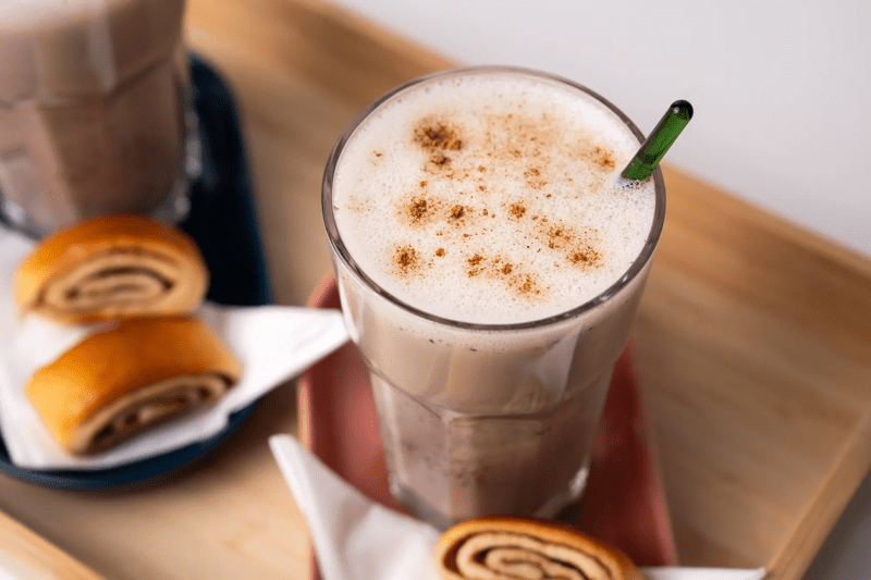 Ein Haferlatte in einem Glas, welches mit Zimtschnecken auf einem Holztablett präsentiert wird.