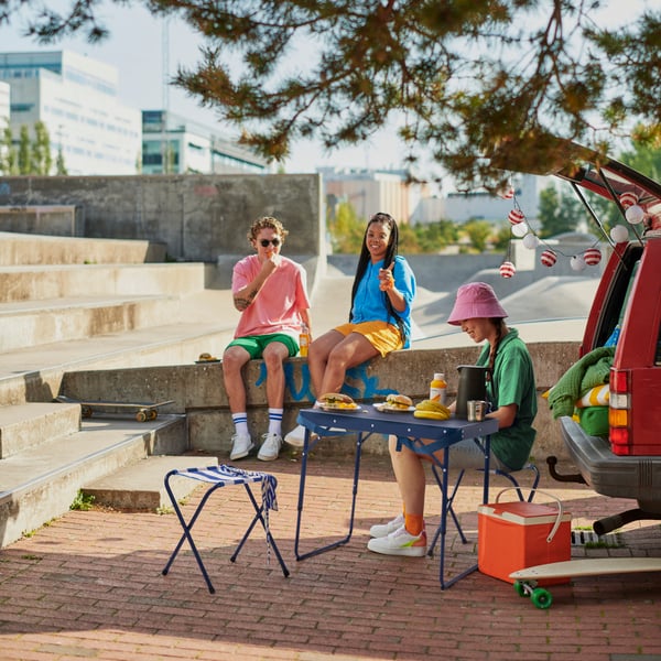 Drei junge Leute genießen ein Picknick mit IKEA Campingmöbeln beim Skatepark – urbaner Lifestyle trifft funktionales Design

