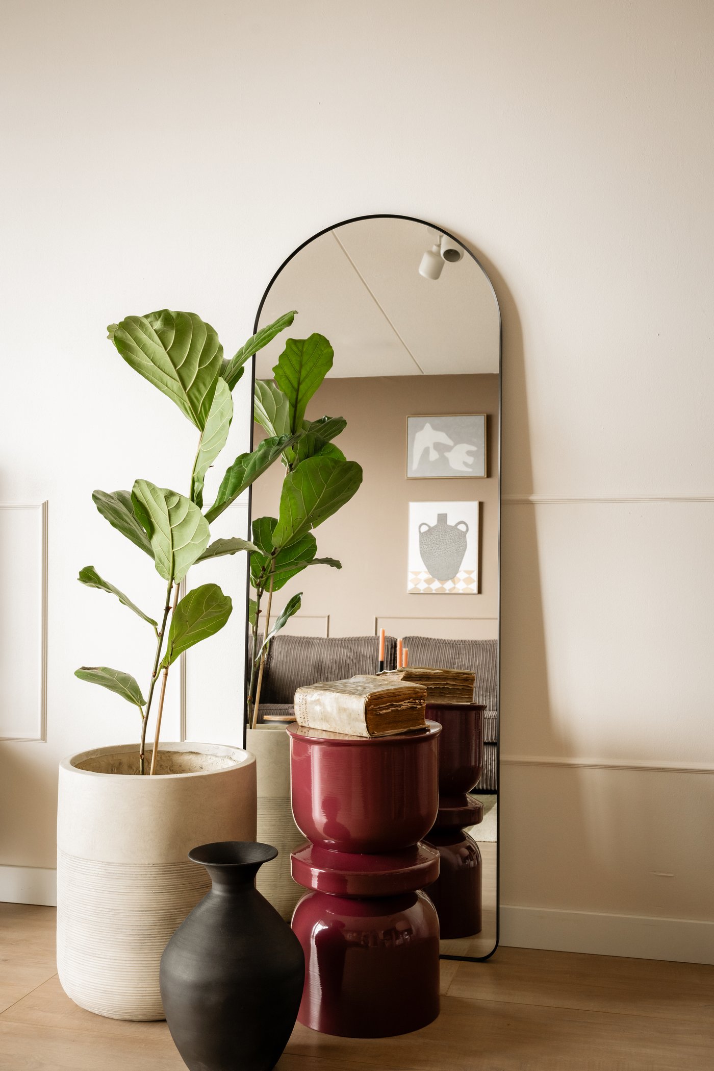 Een beige muur waartegen een grote, afgeronde LINDBYN spiegel staat met zwarte rand. Hiervoor staat een grote plant in pot, een donkerrood bijzettafeltje en een losse donkerbruine vaas.