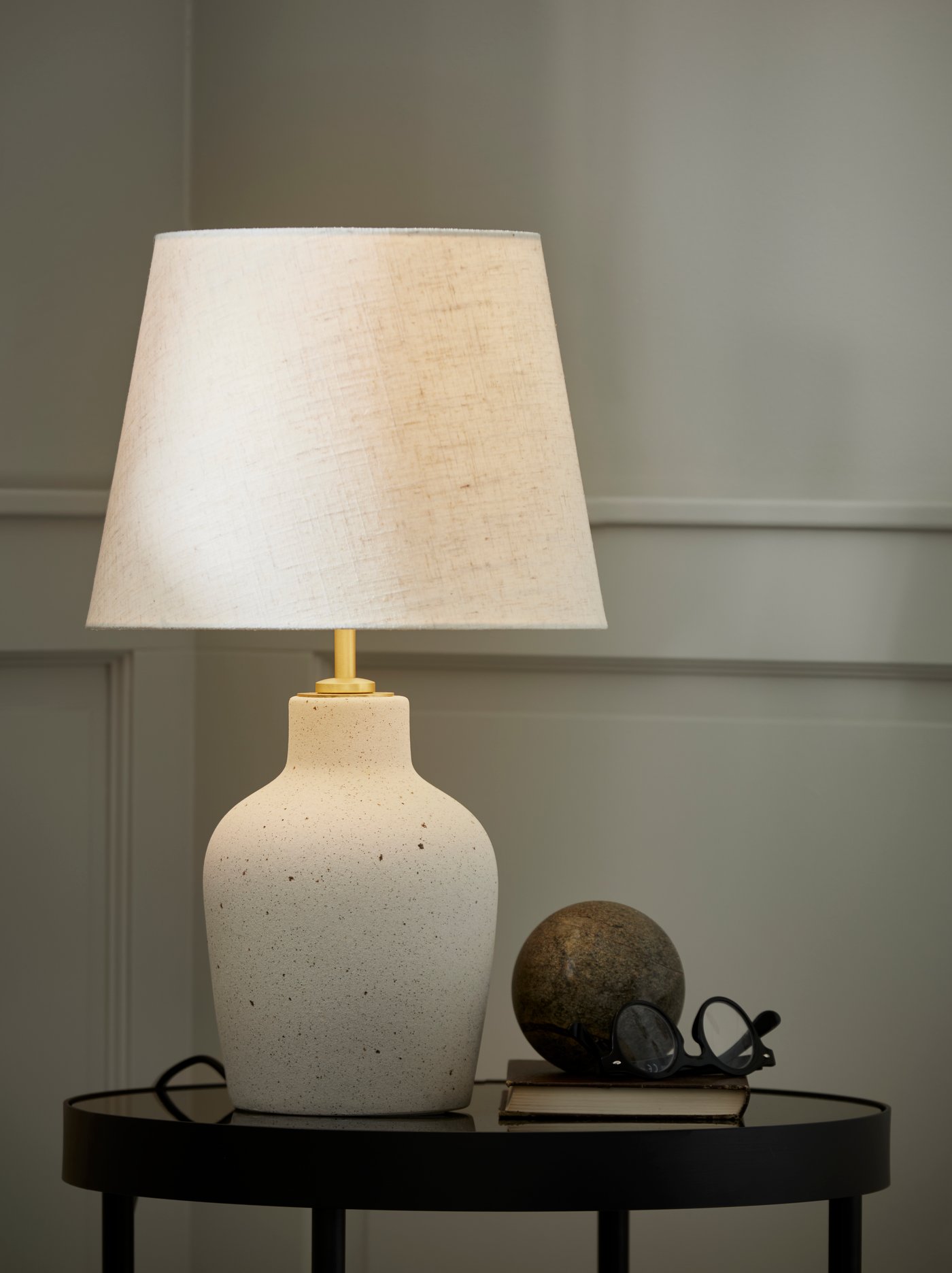 Stolna lampa s bijelim sjenilom na crnom okruglom stolu, uz knjigu, kuglu i naočale.