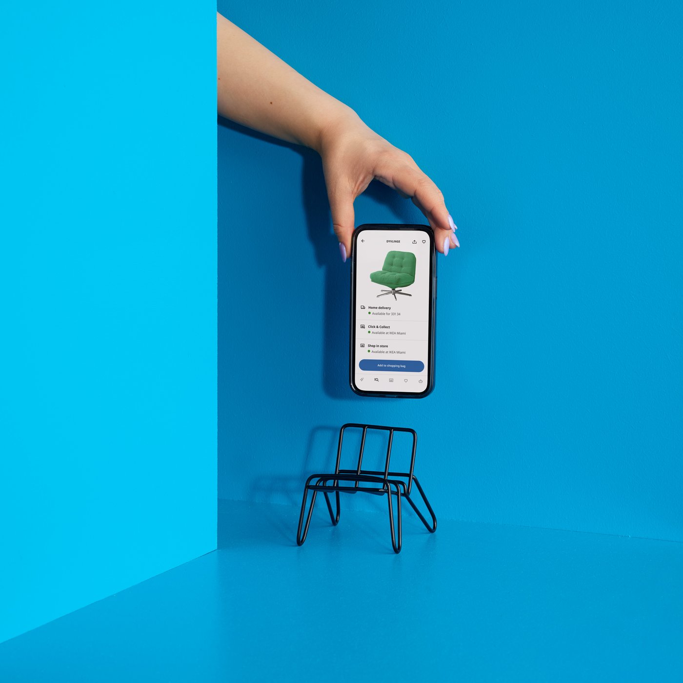 A hand holds a smartphone against a blue wall above a small black chair-shaped phone holder, showing a product page in the IKEA app.