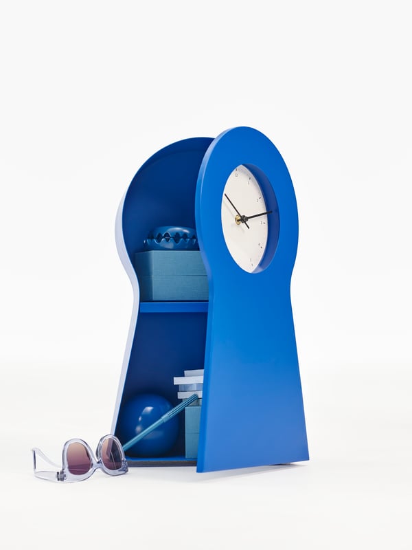 A blue clock in a white space. The clock front is open, showing off the storage shelf inside.