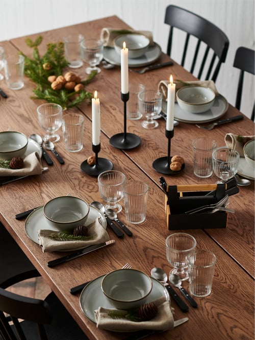 A wooden table set with GLADELIG crockery, glasses, napkins, candles, evergreen branches and nuts scattered.