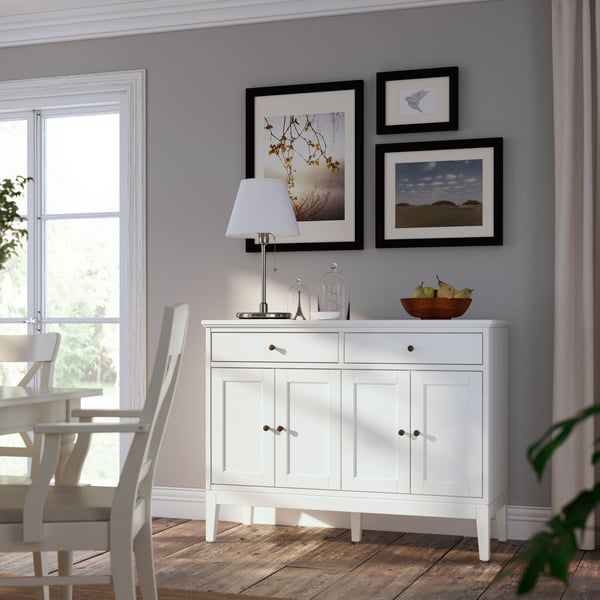 IDANÄS storage cabinets in a living room with an ARSTID table lamp on top of it, below a few KNOPPANG frames with posters. 