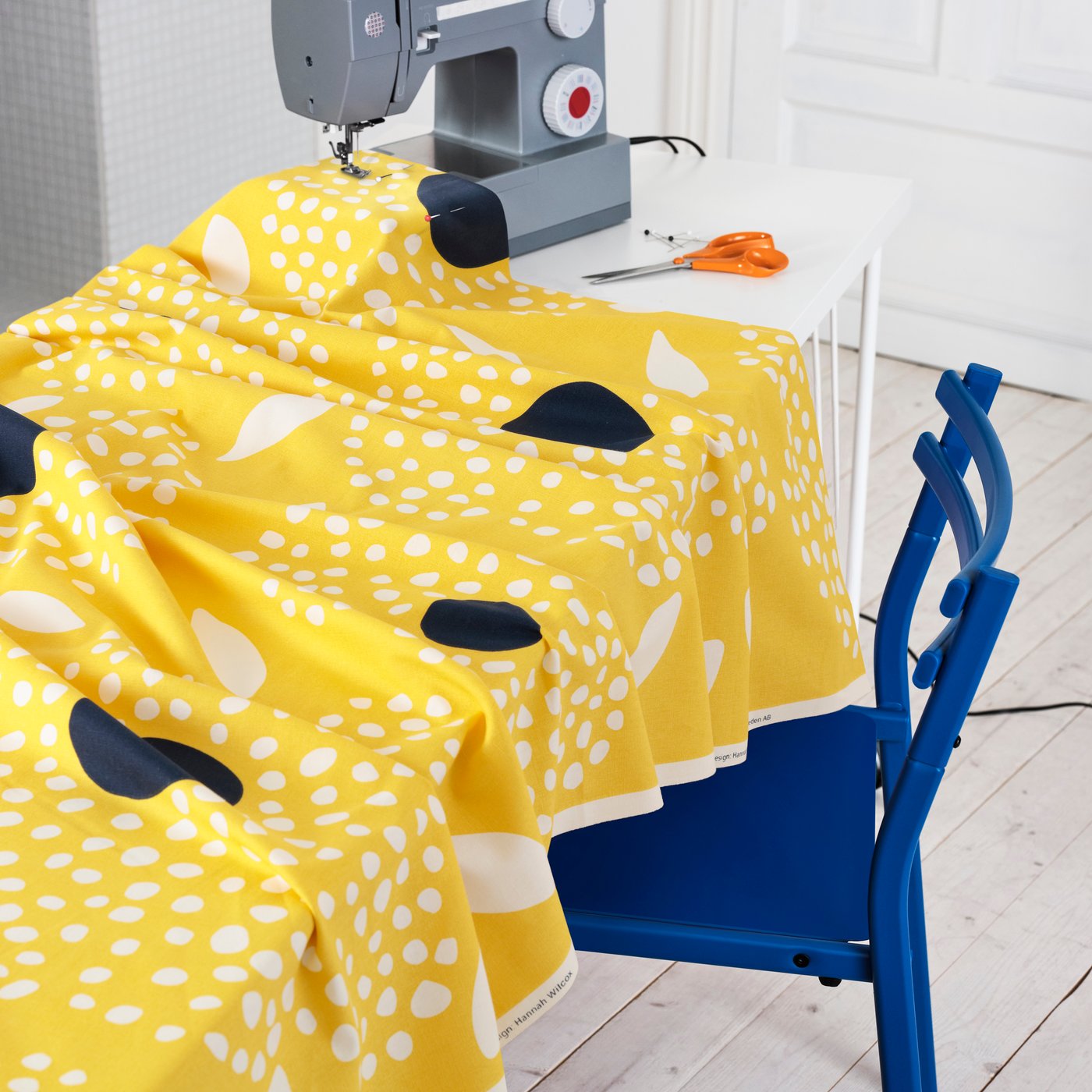 Un tessuto pretagliato BUSKOPUNTIA con motivo giallo, nero, bianco e a pois sul tavolo con una macchina da cucire e una sedia blu.