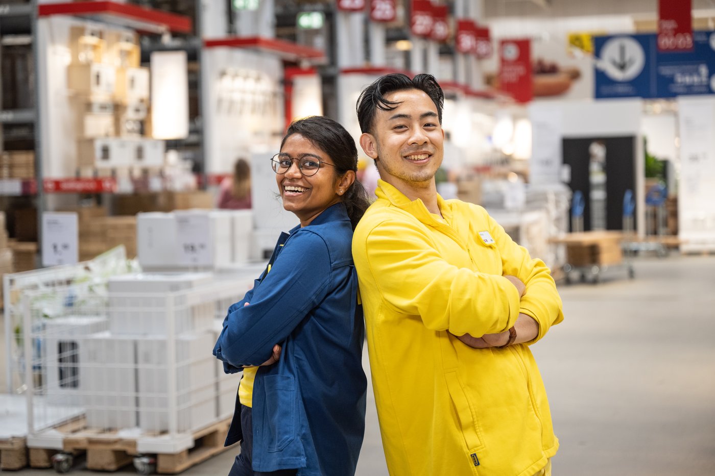 Ein IKEA Mitarbeiter und eine IKEA Mitarbeiterin stehen Rücken an Rücken in er SB-Halle eines Einrichtungshauses