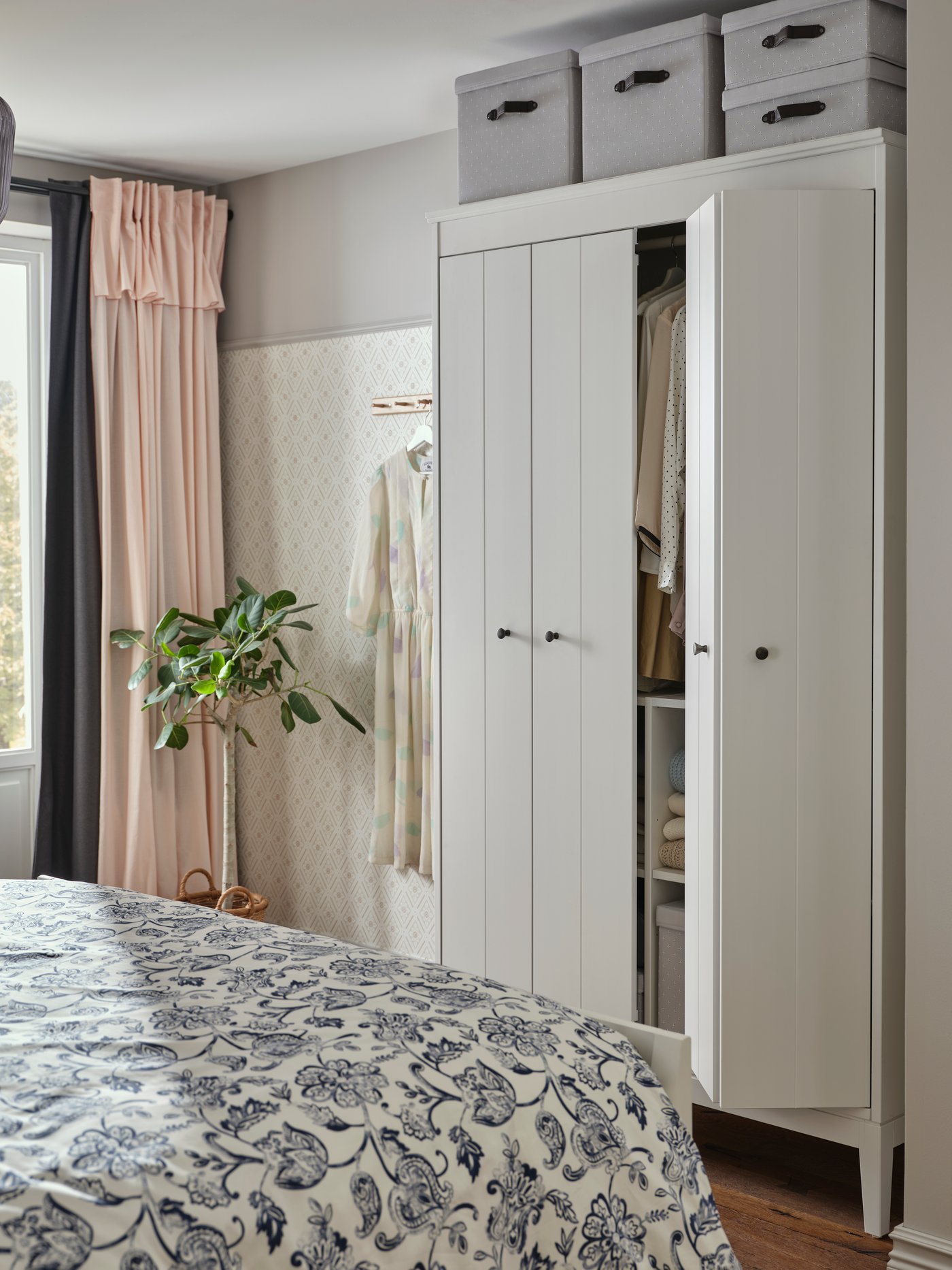 Sovrum med vit IDANÄS garderob där en dörr står på glänt och visar kläder och vikta textilier på hyllor, med mönstrade gardiner och en växt intill fönstret.