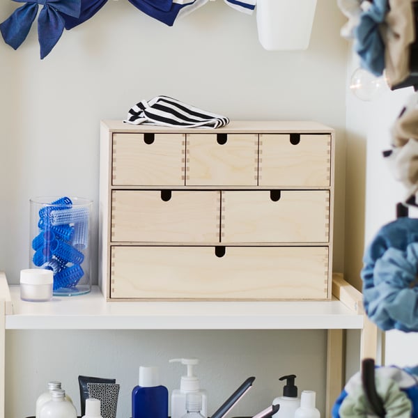 In a bathroom, a MOPPE mini chest of drawer in birch plywood and holding toiletries and accessories, is on a shelving unit.