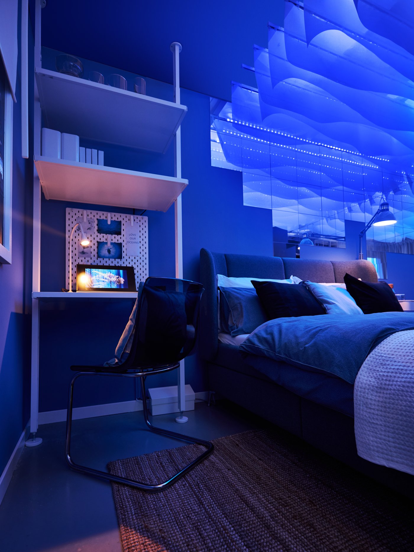 Ett sovrum i blå belysning med en säng, kuddar och filt, samt ett litet skrivbord med hyllor och en stol bredvid.