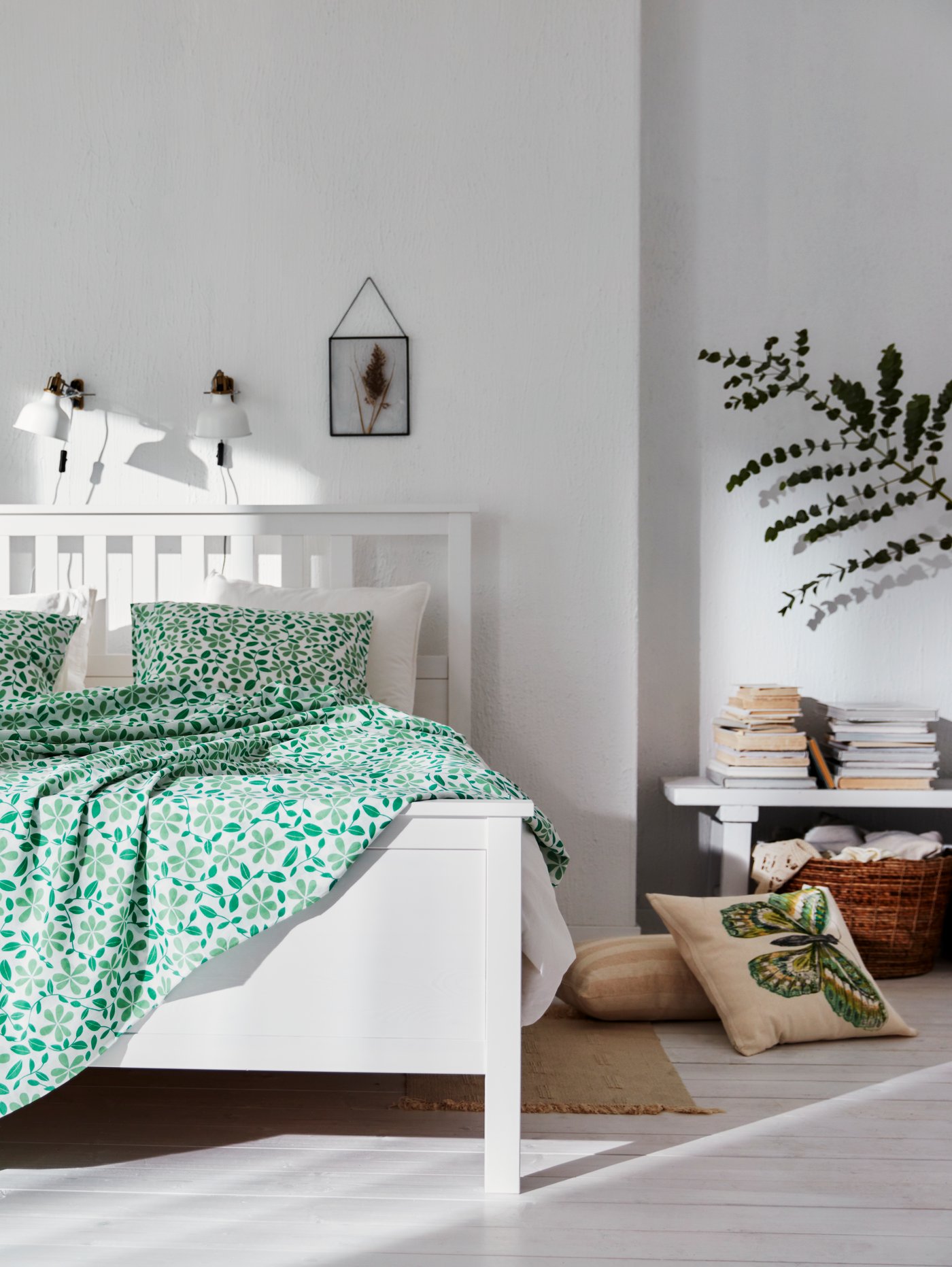 Dormitorio con una estructura de cama, fundas nórdica y de almohada, un foco de pared, un marco gris, una funda de cojín cuadrada, una alfombra, libros y plantas.