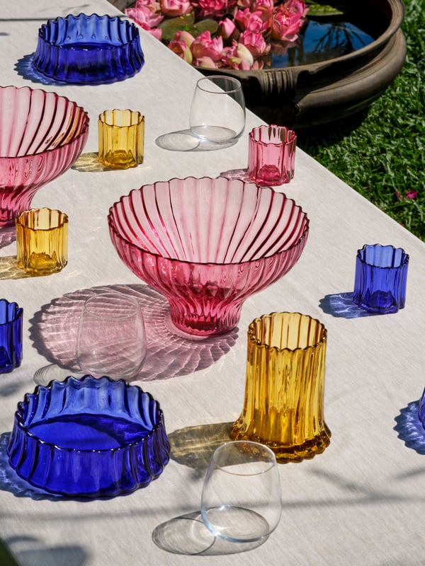 Table full of colourful glass AROMATISK tealight holders, vases and bowls that are reflected in the sunlight