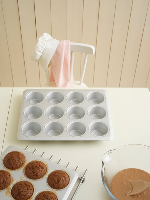 Ujęcie z góry stołu z dwiema foremkami do muffinek, jedną pustą, a drugą z muffinkami na podstawce, miską i krzesłem z ubraniami.