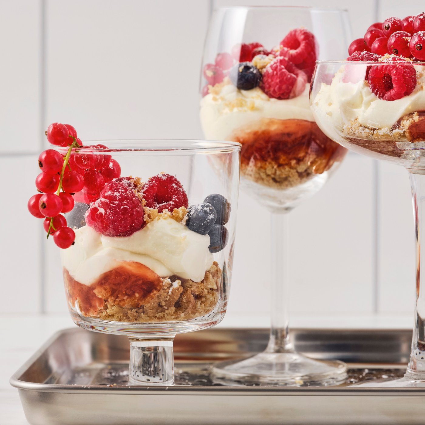 Several glasses of different shapes, with oat biscuit and raspberry jam cheesecake inside, are placed on a metal tray.