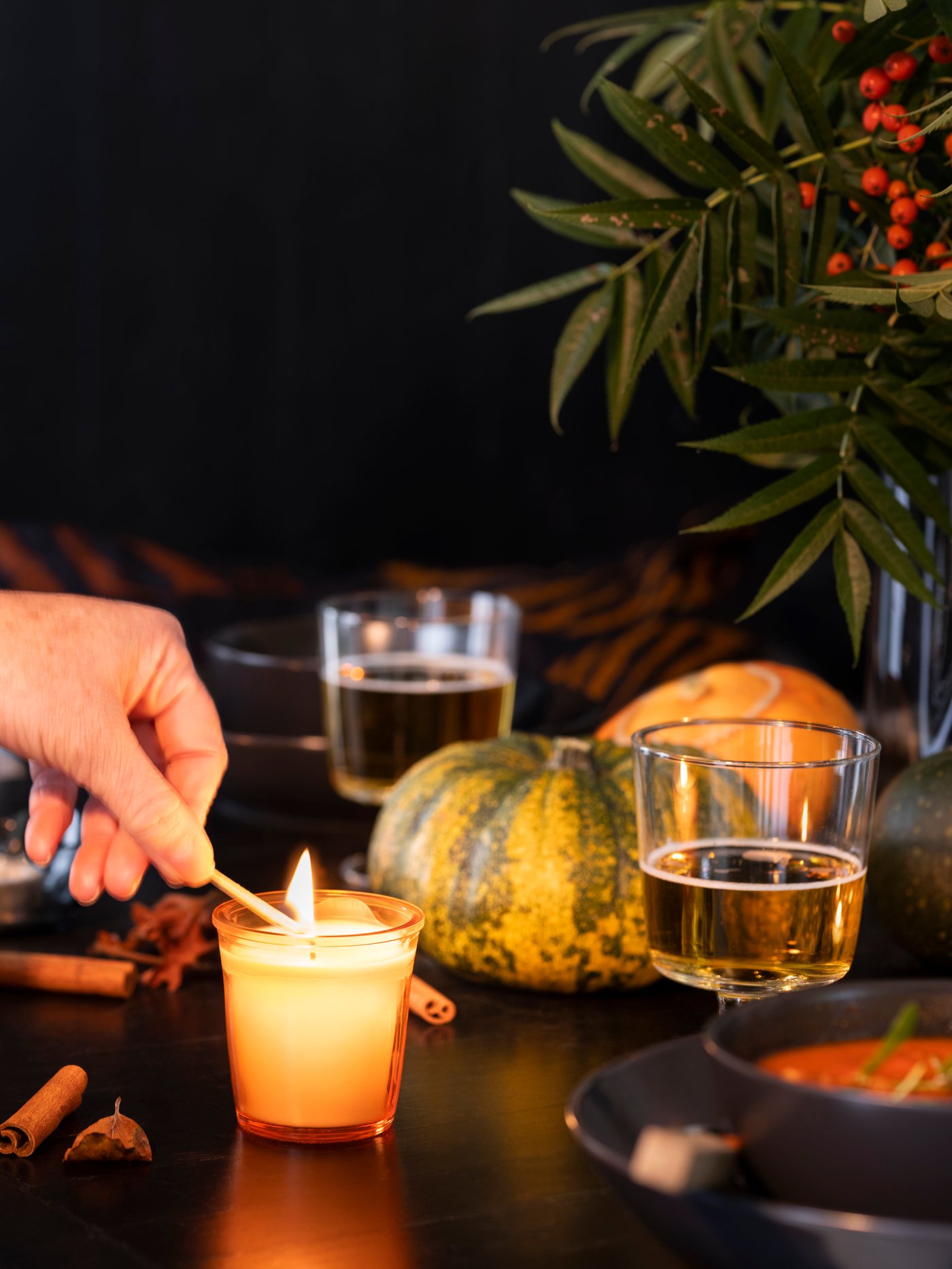 Una mano accende una candela posta su un tavolo. Attorno, decorazioni di zucca tipiche autunnali accanto a bicchieri.