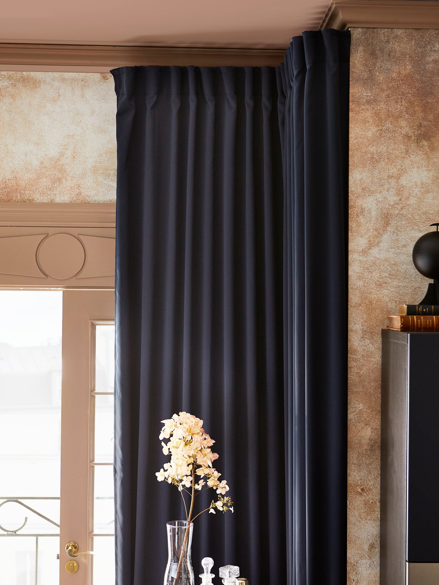 A room with MAJGULL block-out curtains in dark blue, drawn back to show the light, and a sprig of flowers in a glass vase.