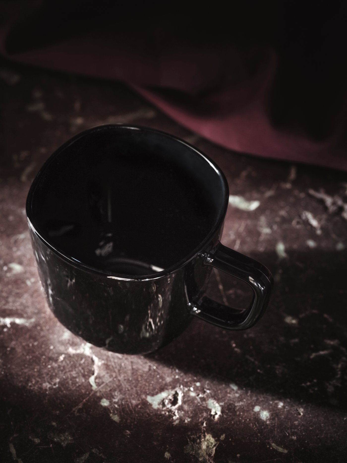 A black glossy BACKIG mug with a handle placed on a dark-red stone marble surface with some fabric in the background.