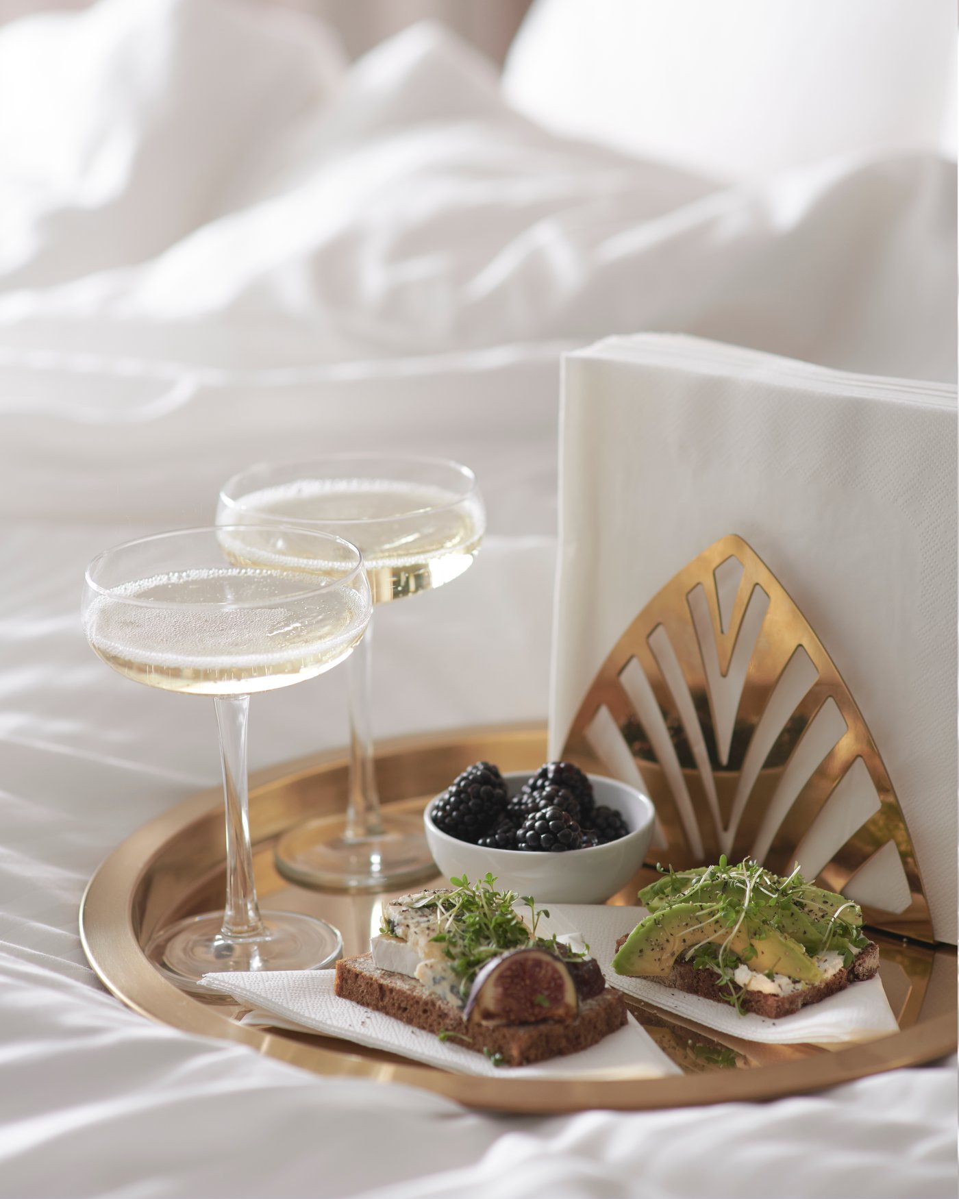 A brass-coloured TILLSTÄLLNING napkin holder on a gold serving tray, on a bed with white bedding.