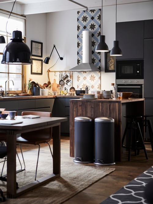 Kitchen area with multiple base cabinets with three drawers, two pedal bins, a table with a chair, pendant lamps, and a rug.