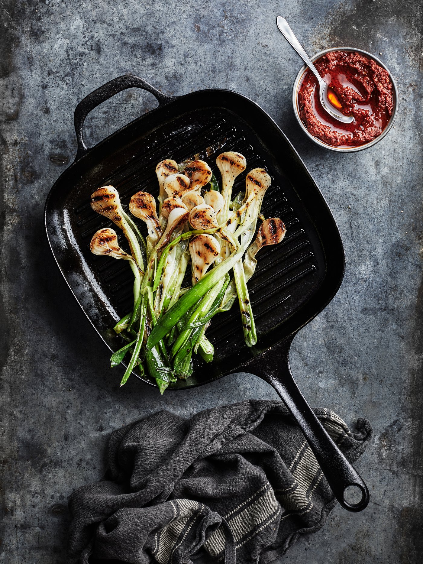 A black grill pan with halved onions being fried inside stands on a stone worktop next to a bowl of red sauce.