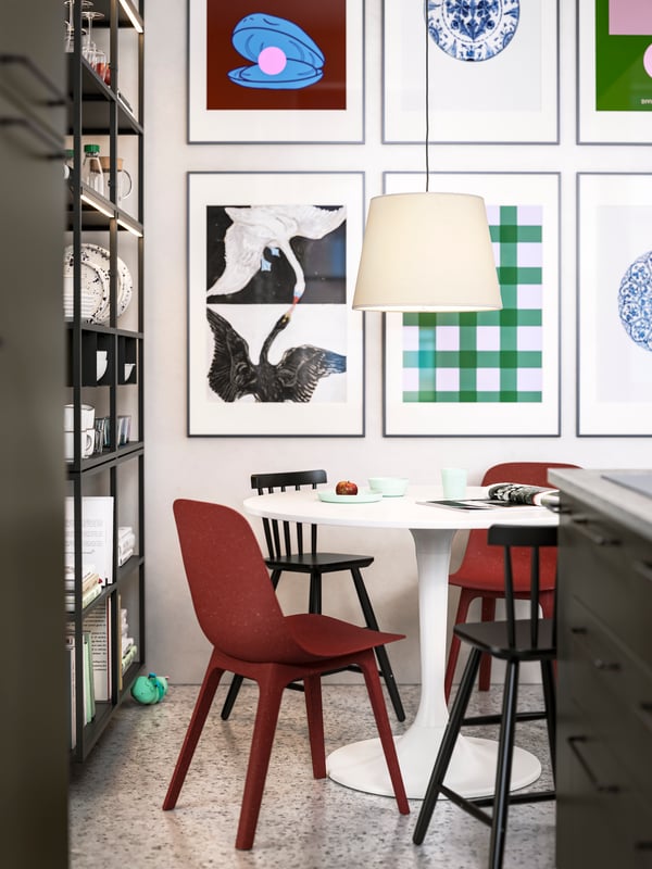 A dining area with a round white table, two red ODGER chairs, two black junior chairs and a pendant lamp, plus framed art.