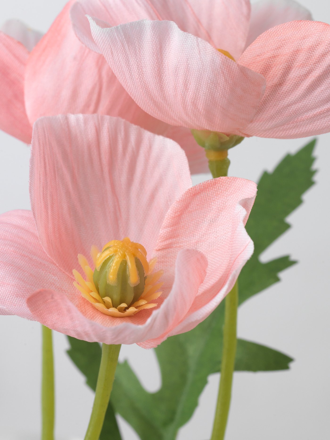 Nahaufnahme einer künstlichen rosa Mohnblume mit gelber Mitte und grünen Blättern, aus der SMYCKA-Serie, vor weißem Hintergrund.