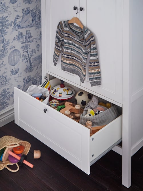 A traditional white SUNDVIK wardrobe with a low open drawer holding toys and a child's sweater hanging above on a door knob.