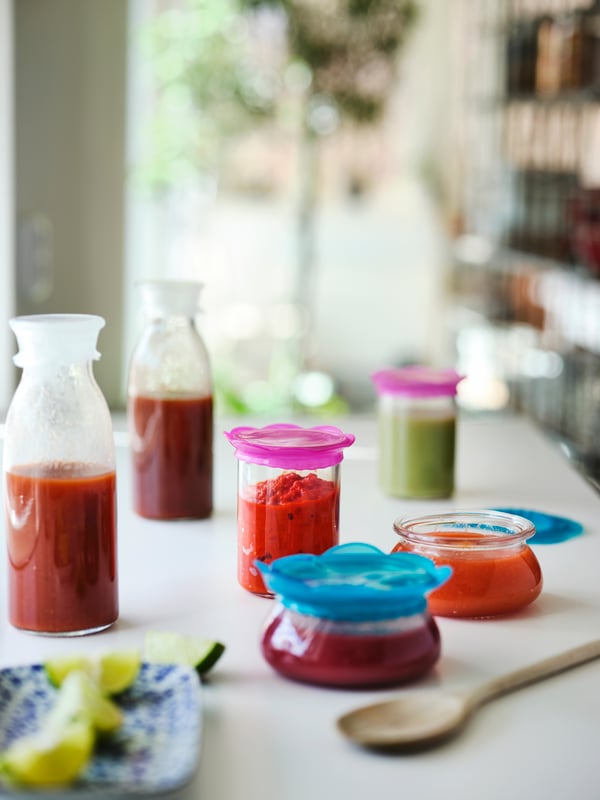 Transparent turquoise and pink silicone ÖVERMÄTT food covers placed on various sauce jars, set on a white countertop.