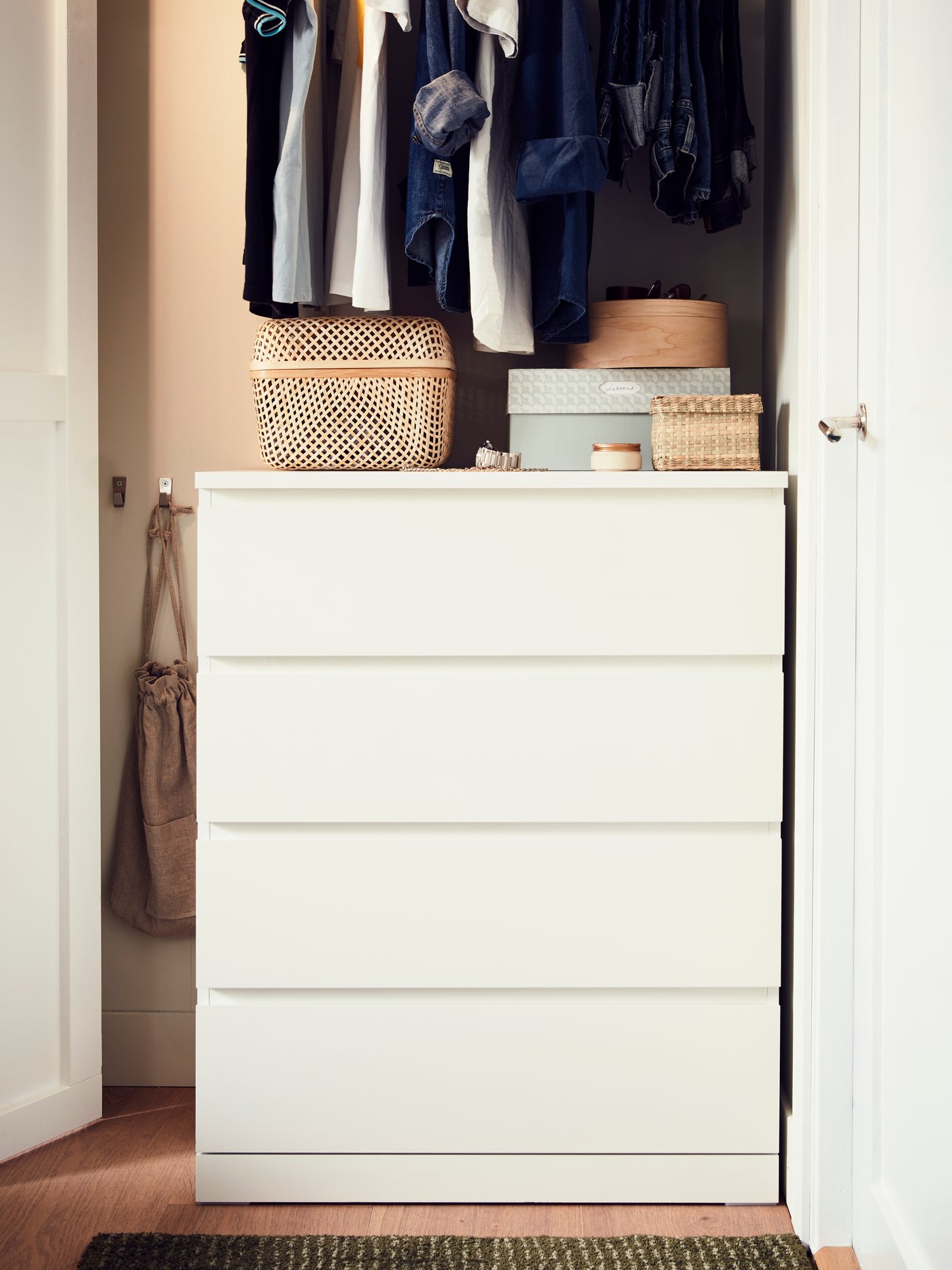 Bijela komoda s četiri ladice ispod viseće odjeće u ormaru. Na vrhu su pletene košare i kutije, a s lijeve strane police s posloženom odjećom i košarama.