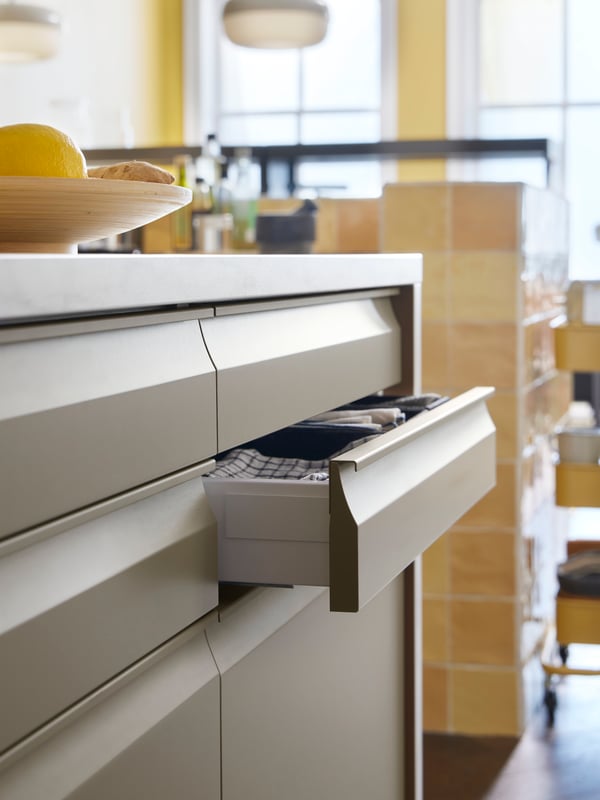 UPPLÖV drawer fronts in matte dark beige, with a drawer open with a tea towel in it, and BLÅSVERK pendant lamps hanging.