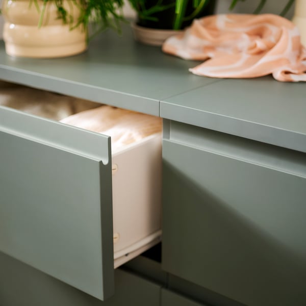 Two gray-green STORKLINTA chests with 3 drawers side by side with the top drawer of one open, with items of clothing inside.