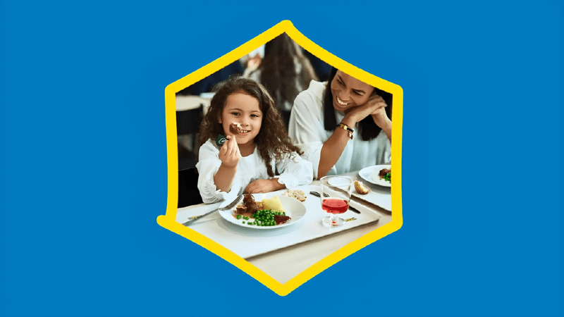 Una bambina è seduta ad un ristorante e sta mangiando un piatto con delle polpette. Accanto a lei una donna la guarda sorridendo.