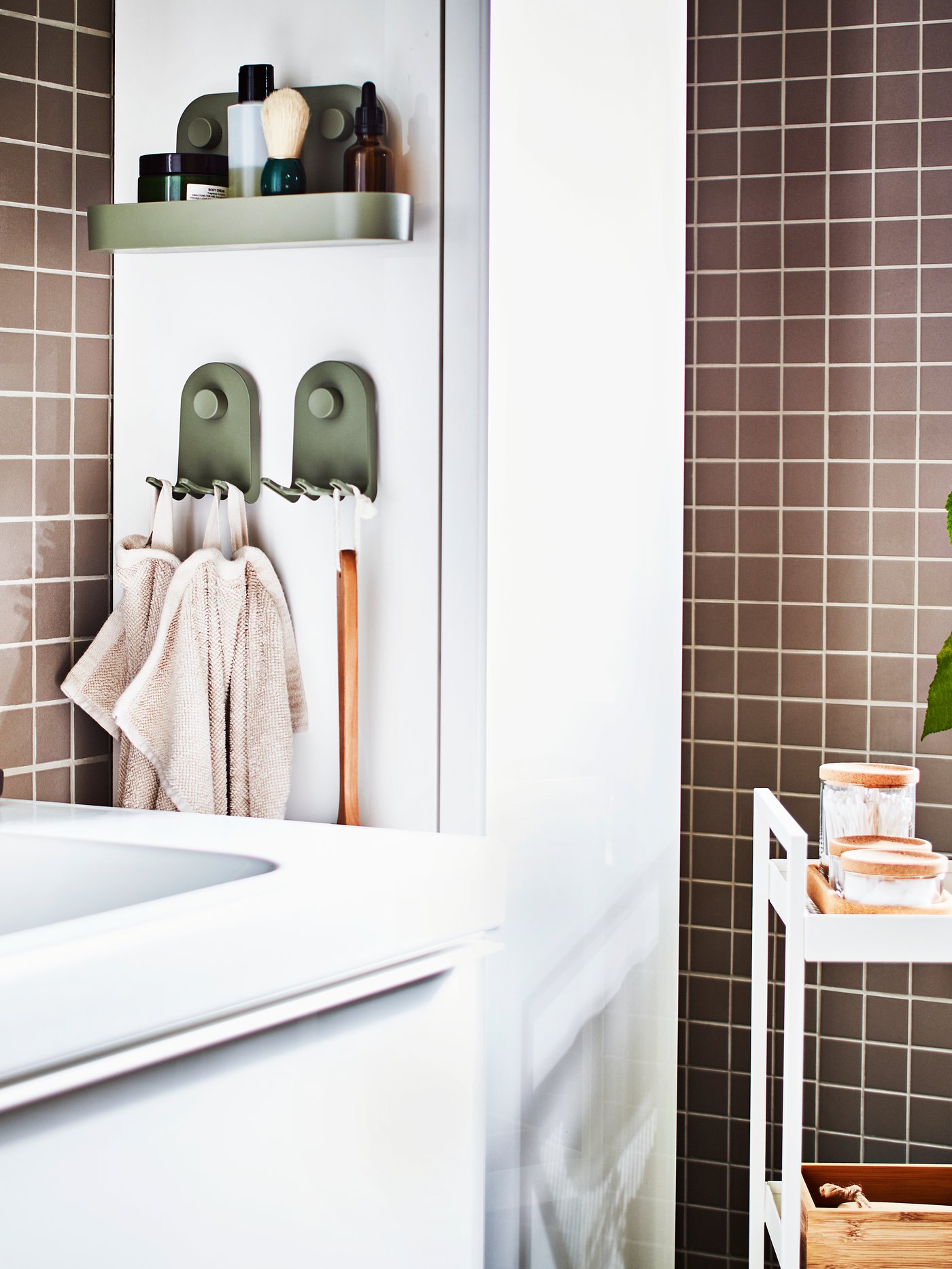 Ett gråkaklat badrum med ett vitt tvättställ, ett vitt högskåp, en vit rullvagn med brickor och burkar samt en krukväxt.