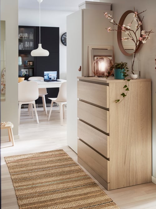 Hol cu lada cu patru sertare, oglinda rotunda pe perete, lampa pandantiv, scaune, masa, carti, obiecte decorative, covor, taburet.