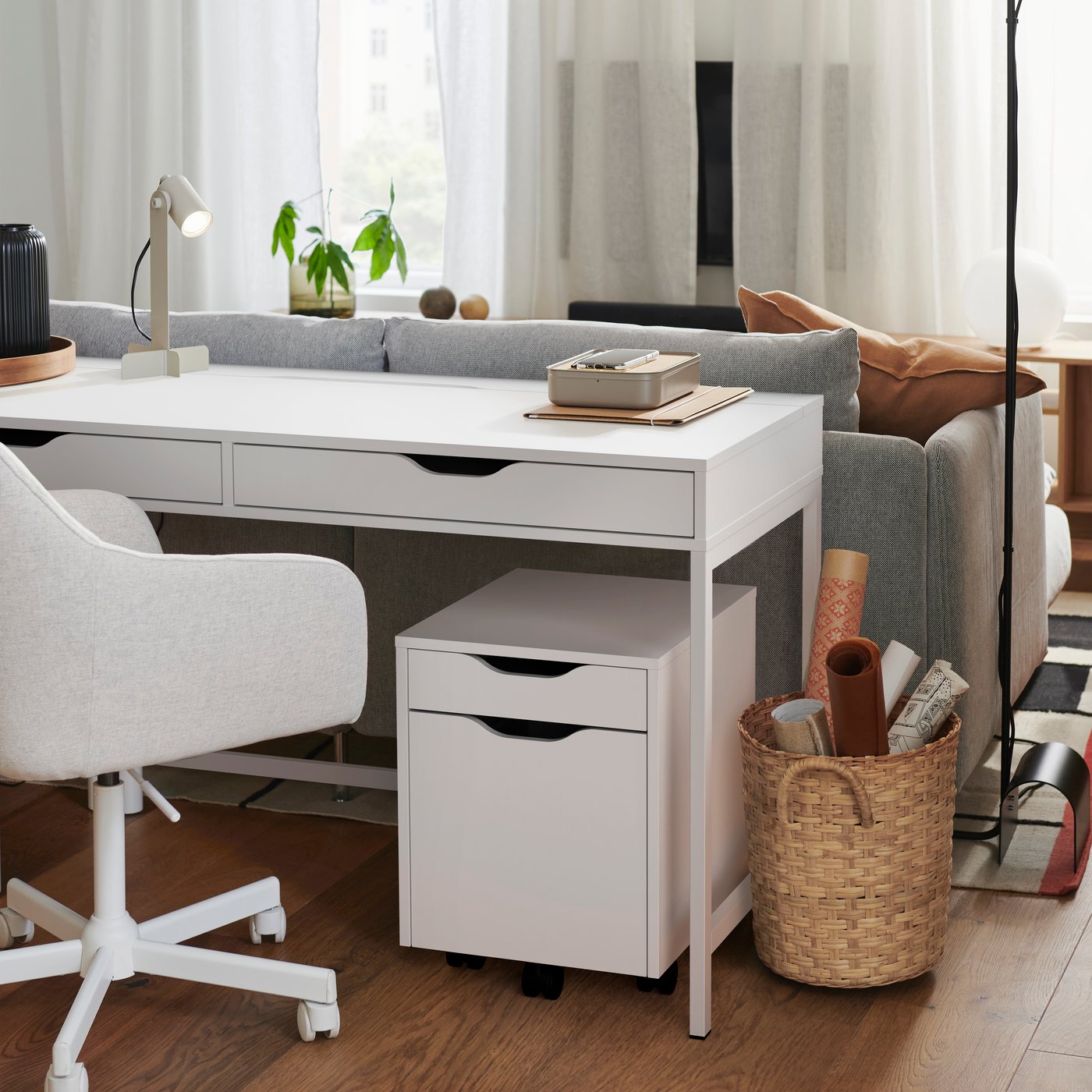 Ett vitt ALEX skrivbord med en ljuspeige skrivbordsstol placerade bakom eb ljusgrå soffa i ett vardagsrum. 