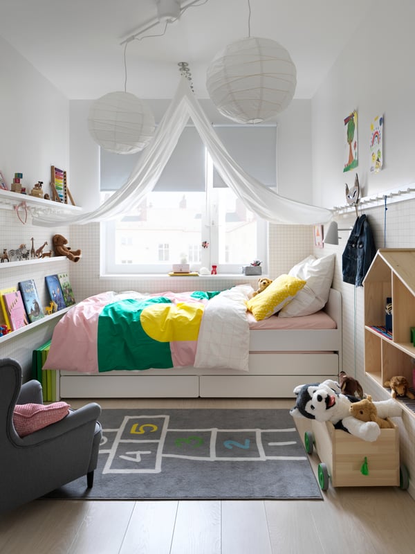 Vista general de una pieza infantil con una cama SLÄKT con cajones inferiores ubicada junto a una ventana. Frente a la cama hay repisas con libros y juguetes, y a un costado se ve una casa de muñecas de madera y una caja para juguetes FLISAT con peluches. 