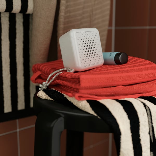 A VAPPEBY portable bluetooth speaker in white placed on folded rust-coloured towels on a black stool in a bathroom.