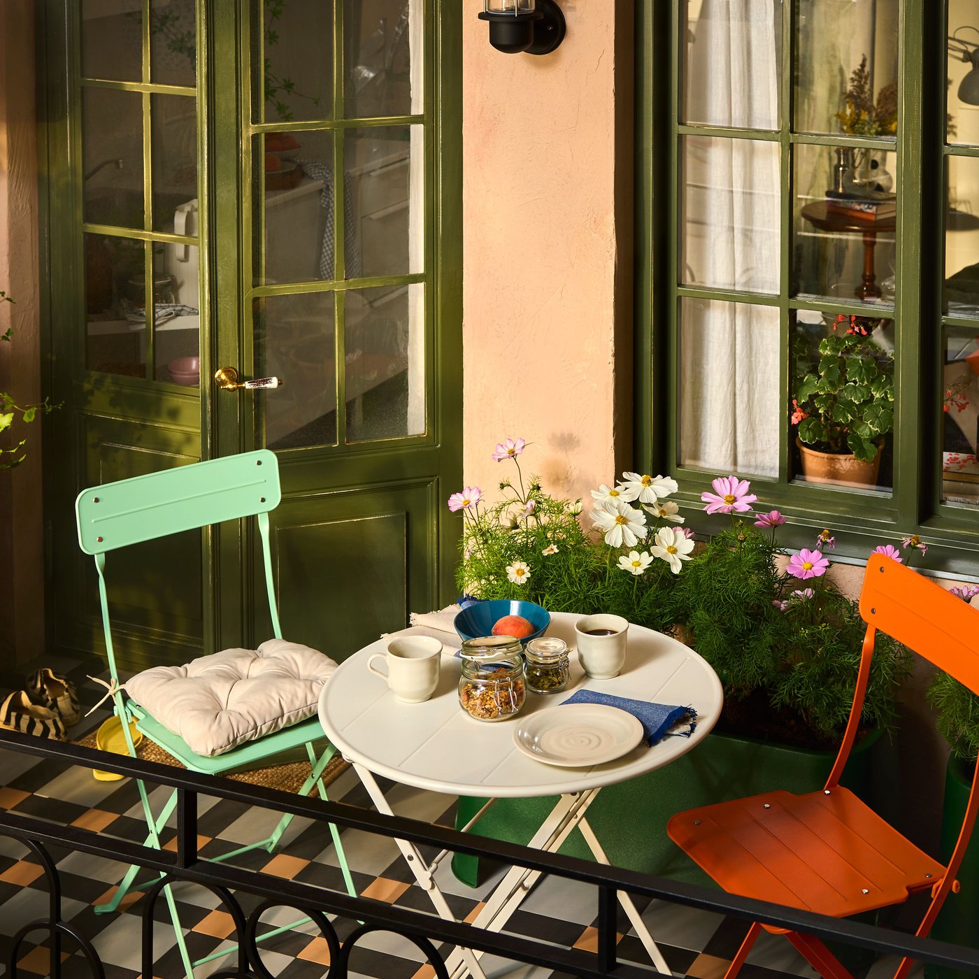 A city balcony with SUNDSÖ outdoor furniture in playful colours surrounded by plants, flowers and cabinets for storage.
