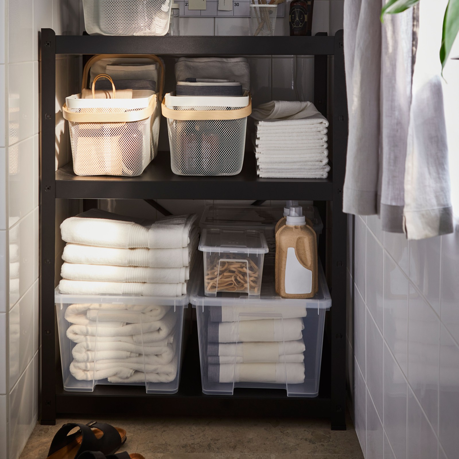 A bathroom interior with a black BROR shelving unit storing clear SAMLA boxes and neatly folded towels on three shelves.