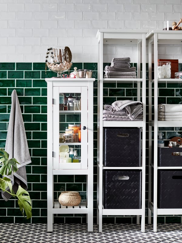 Una vetrina HEMNES e scaffali bianchi con contenitori e accessori per il bagno, tra cui diversi prodotti tessili. La parete ha piastrelle bianche e verde scuro.