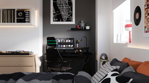 Chambre avec lit au premier plan et bureau contre un mur peint en noir à l’arrière-plan, avec un ordinateur et un fauteuil gamer.