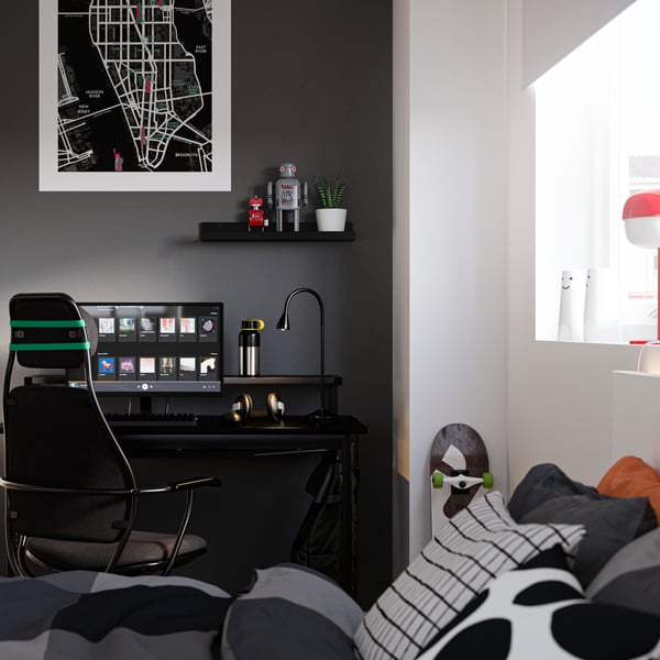 Bedroom with a bed in the foreground and a desk by a wall painted black in the back, with a computer and gaming chair.