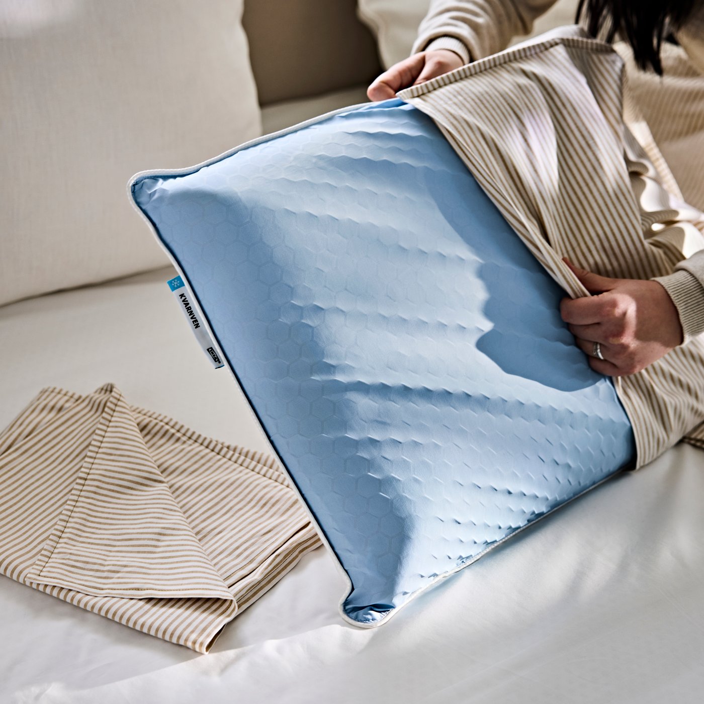 A person is putting on a striped LAGERPOPPEL pillow case on a KVARNVEN ergonomic pillow.