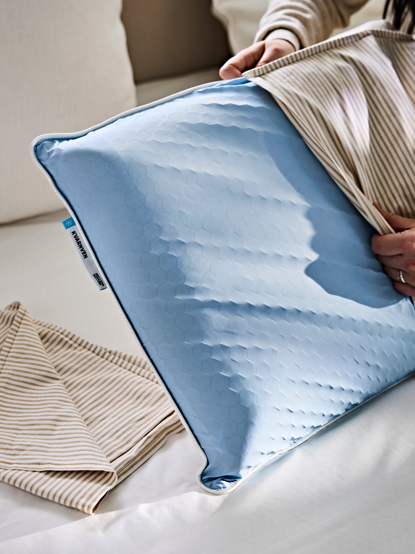 Een persoon doet een gestreepte LAGERPOPPEL kussensloop om een KVARNVEN ergonomisch kussen.