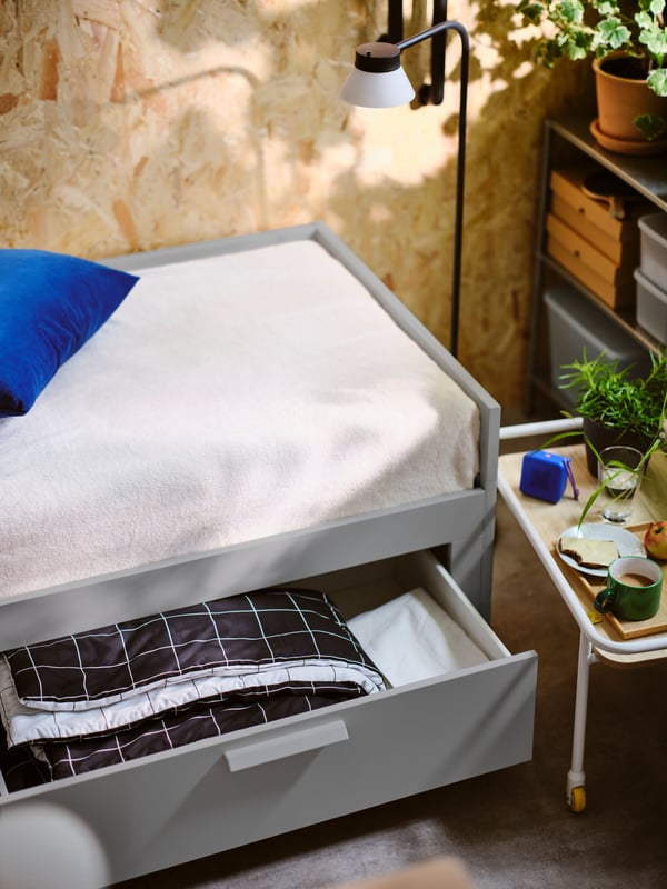 An open drawer of a grey BRIMNES day bed with 2 drawers showing a folded black/white VITKLÖVER comforter set inside.