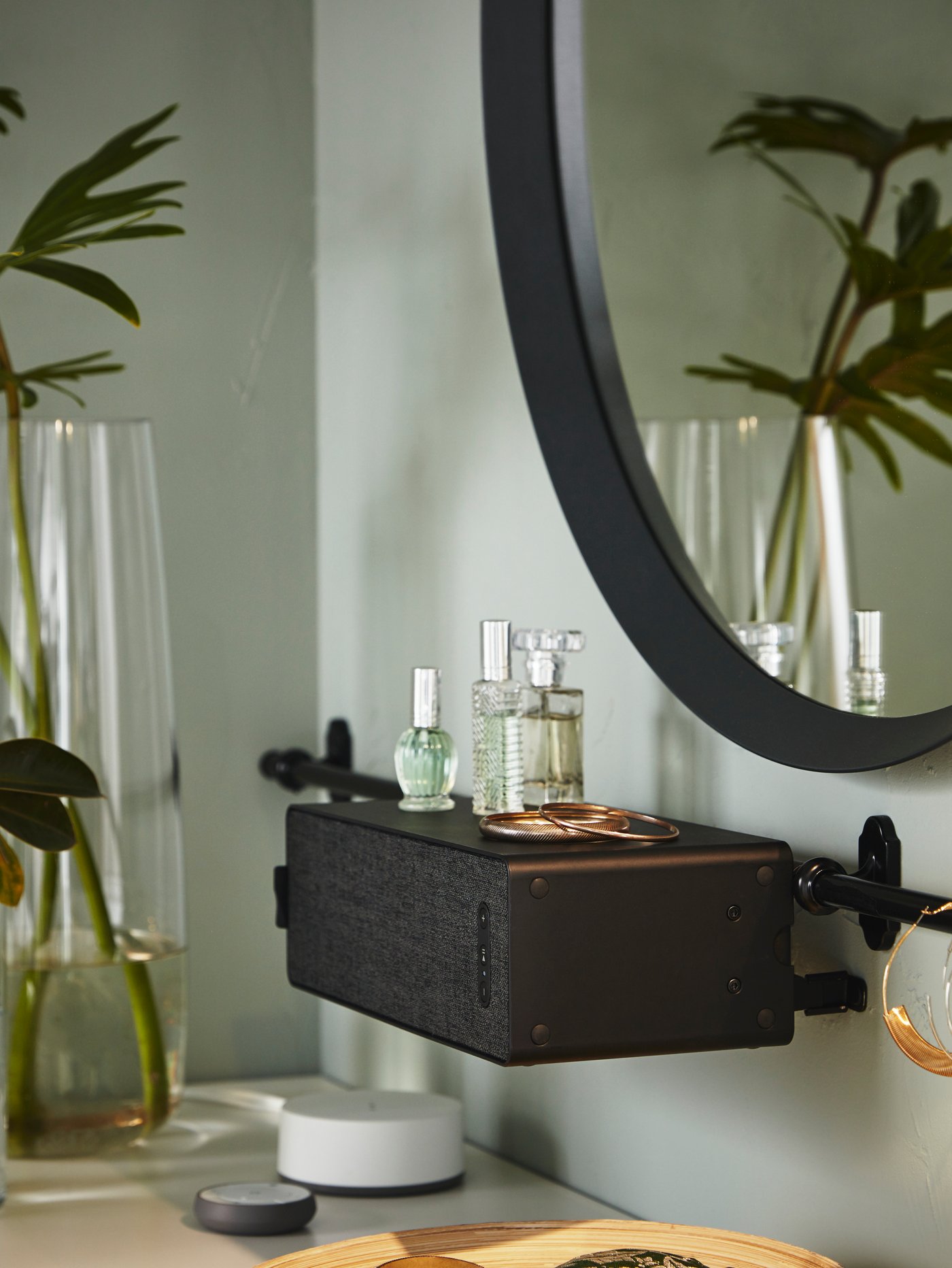 Policový reproduktor SYMFONISK je upevněn ke stěně, stojí na něm parfémy a doplňky, nad ním visí zrcadlo LANGESUND.