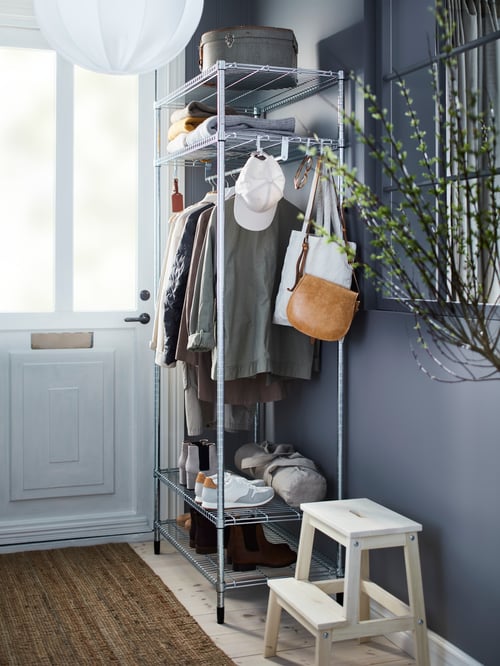 Hallway with a shelving unit with clothes rail, with footwear, clothes on hangers, and bags. A stool is next to the unit.