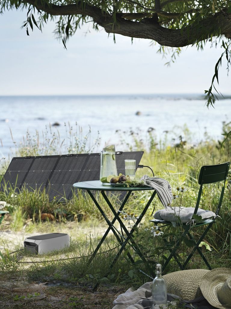 In een rustige buitenomgeving staat een IKEA-tuinset naast een zonnepaneel, vlak bij het water en in de schaduw van een boom.