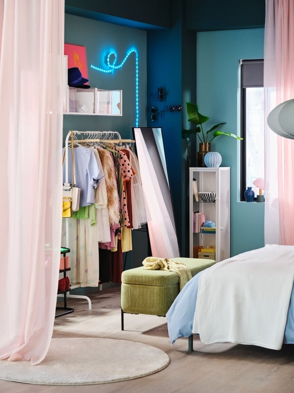 A green VALTORP bench with storage at the end of a bed, in front of a clothes rail and a sheer pink STENFRÖ curtain.