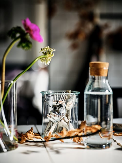 A close up shot of a dining room featuring a carafe with a stopper, a jar with a 16-piece cutlery set inside, and a vase.